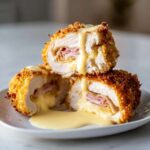 Two halves of crispy, breaded chicken cordon bleu stacked, revealing melted cheese and ham inside, drizzled with sauce.