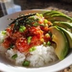 A vibrant bowl of spicy tuna rice topped with sesame seeds, nori, and green onions, served with sliced avocado.
