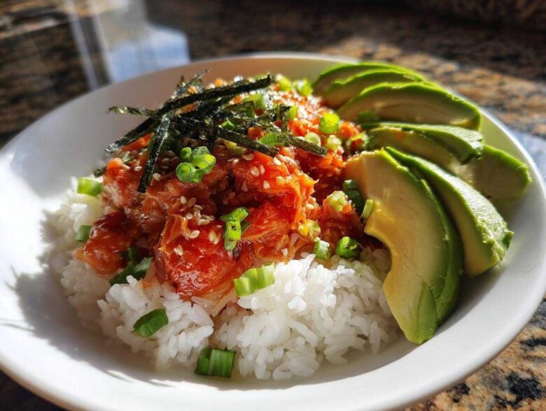 A vibrant bowl of spicy tuna rice topped with sesame seeds, nori, and green onions, served with sliced avocado.
