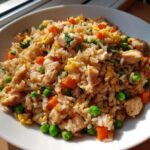 Close-up of a bowl of amazing chicken fried rice mixed with diced chicken, bright green peas, and orange carrots.