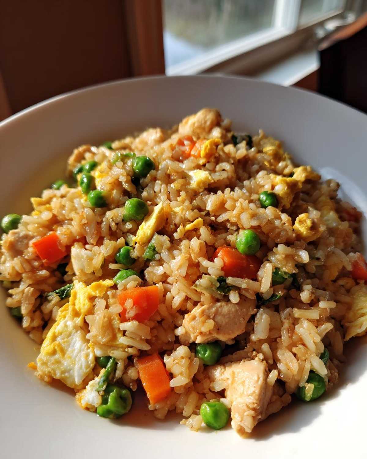 A close-up, brightly lit serving of amazing chicken fried rice with peas, carrots, and scrambled egg pieces.