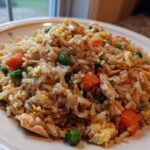 A close-up of a serving of amazing chicken fried rice featuring rice, chunks of chicken, bright green peas, and diced carrots on a white plate.