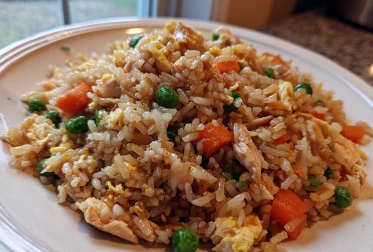 A close-up of a serving of amazing chicken fried rice featuring rice, chunks of chicken, bright green peas, and diced carrots on a white plate.
