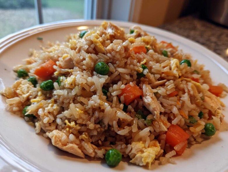 A close-up of a serving of amazing chicken fried rice featuring rice, chunks of chicken, bright green peas, and diced carrots on a white plate.