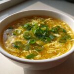 A white bowl filled with golden yellow egg drop soup, featuring wispy cooked egg ribbons and topped generously with fresh green chopped scallions.