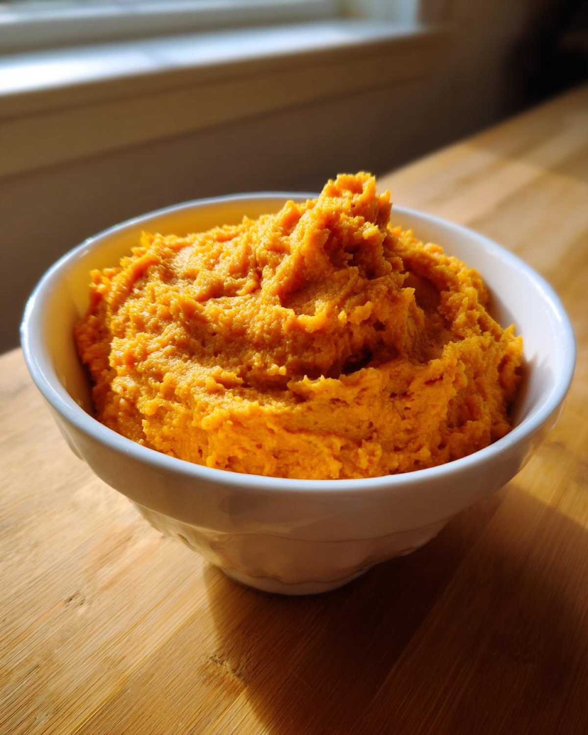 A white bowl filled high with bright orange, fluffy mashed sweet potatoes, sitting on a wooden surface.