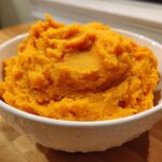 A close-up of a white bowl filled with vibrant, fluffy mashed sweet potatoes.