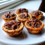 Six freshly baked pecan tassies with gooey filling and pecan halves arranged on a white plate.