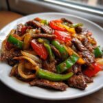 Close-up of glossy, saucy pepper steak stir-fry with colorful bell peppers and onions on a white plate.