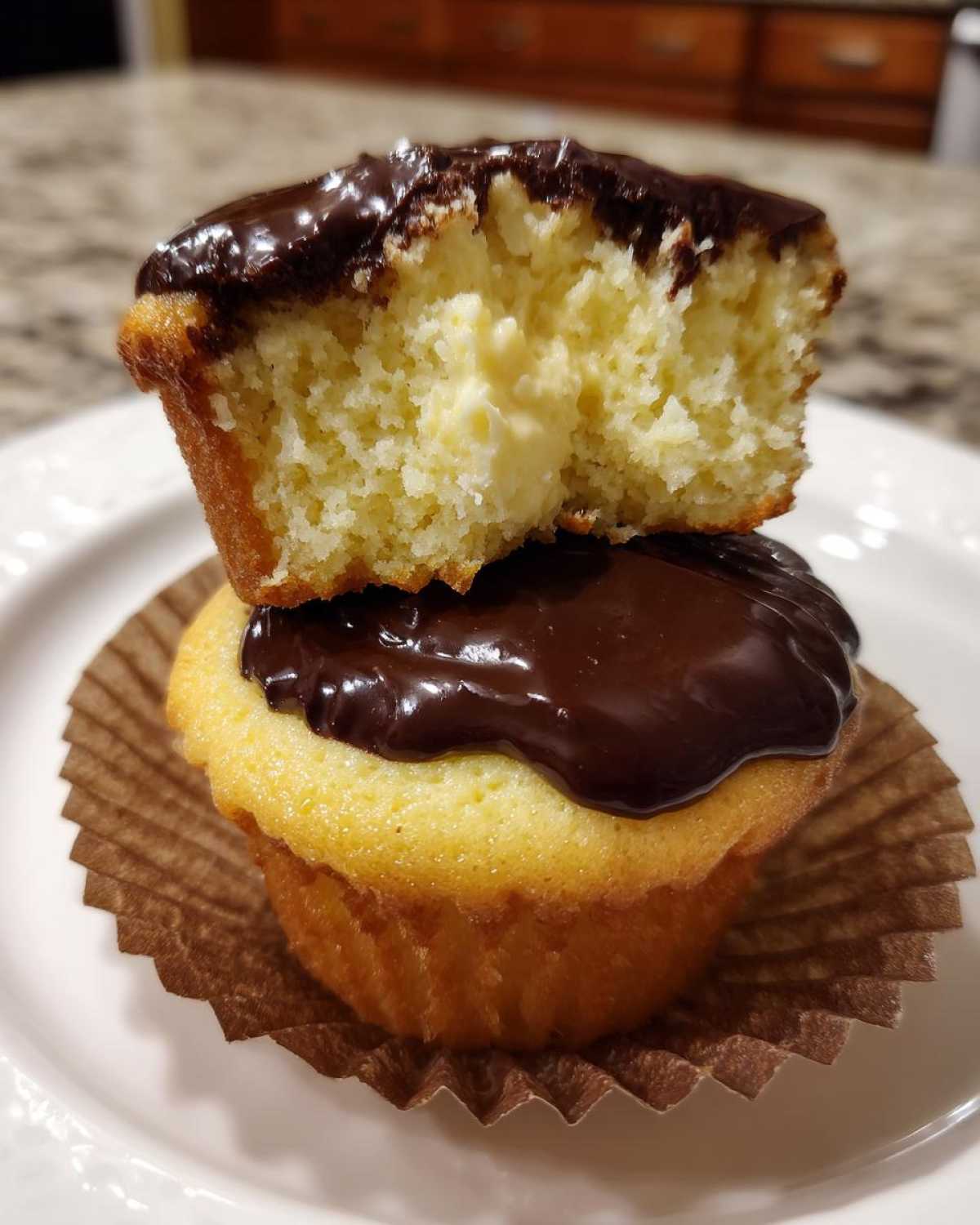 A boston cream pie cupcake cut in half showing the vanilla cake, pastry cream filling, and chocolate glaze.