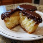 Close-up of a boston cream pie cupcake cut in half showing vanilla cake, pastry cream filling, and chocolate glaze.