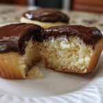 Close-up of a boston cream pie cupcake cut in half showing vanilla cake, cream filling, and chocolate glaze.