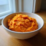 A white bowl filled with bright orange, fluffy mashed sweet potatoes sitting on a wooden surface.