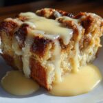 A close-up of a square serving of baked bread pudding recipe, drizzled heavily with creamy vanilla sauce.