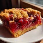 Close-up of a square slice of rich, bright red cherry dump cake with a crumbly topping on a white plate.