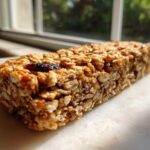 A single, chewy homemade granola bar made with oats and dried fruit, sitting on a white surface near a window.