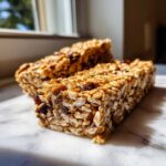 Two chewy homemade granola bars packed with oats and dried fruit sitting on a white marble surface.