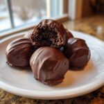 A stack of decadent, chocolate-covered Oreo balls on a white plate, one bitten open showing the dark center.