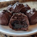 A plate of rich, chocolate-dipped Oreo balls, one cut in half showing the dark, dense interior.