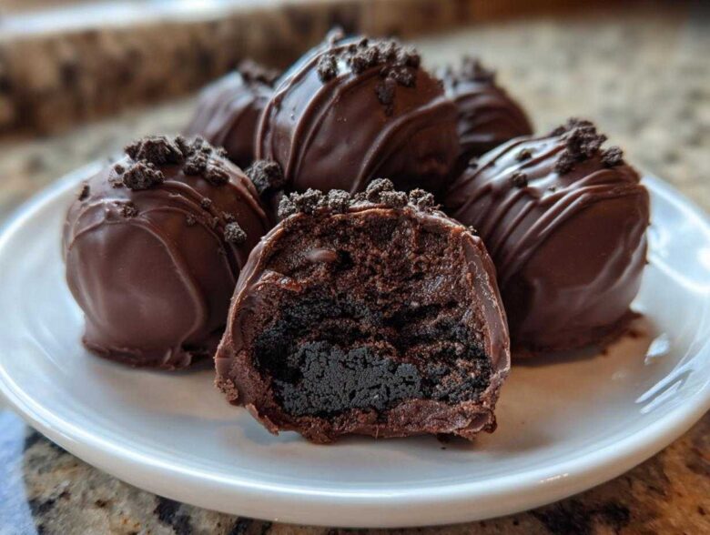 A plate of rich, chocolate-dipped Oreo balls, one cut in half showing the dark, dense interior.