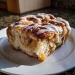 A close-up of a square slice of cinnamon roll apple pie, drizzled with white icing, sitting on a white plate.