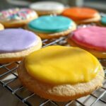 Close-up of sugar cookies with vibrant, smooth sugar cookie icing in yellow, purple, and blue colors drying on a wire rack.