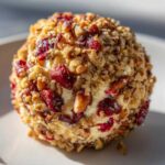 A close-up, appetizing shot of a round cranberry pecan cheese ball coated heavily in chopped pecans and dried cranberries.