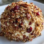 A close-up shot of a perfectly formed cranberry pecan cheese ball coated in chopped nuts and dried cranberries.