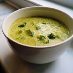 A close-up of a white bowl filled with thick, creamy broccoli cheddar soup, featuring visible florets.