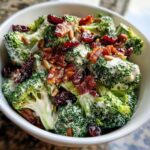 Close-up of creamy broccoli salad topped with bacon bits, dried cranberries, and sunflower seeds.