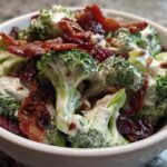 A close-up view of a white bowl filled with creamy broccoli salad, topped with crispy bacon, dried cranberries, and sunflower seeds.