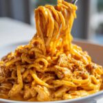 Close-up of creamy, orange-hued cajun chicken pasta being lifted high on a fork from a white bowl.