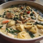 Close-up of a white bowl filled with creamy chicken tortellini soup, featuring tortellini, chicken chunks, spinach, and carrots.