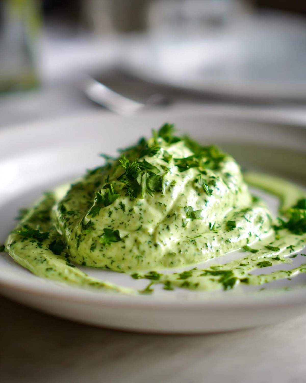 A thick dollop of bright green, creamy cilantro lime sauce garnished with fresh chopped herbs on a white plate.