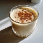 A glass of creamy coquito topped generously with ground cinnamon powder, sitting in sunlight.