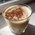 A close-up of a glass filled with creamy coquito, topped with a dusting of ground cinnamon.