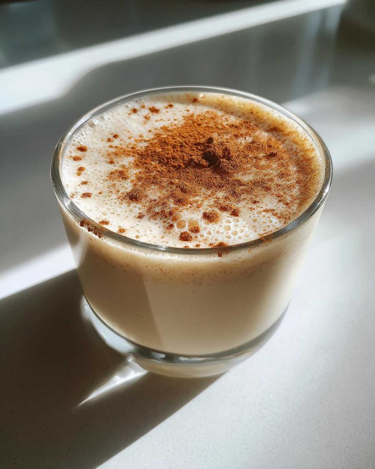 A glass of creamy coquito topped generously with ground cinnamon, sitting on a bright white surface.