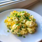 A serving of creamy, yellow egg salad recipe garnished generously with chopped green chives on a white plate.