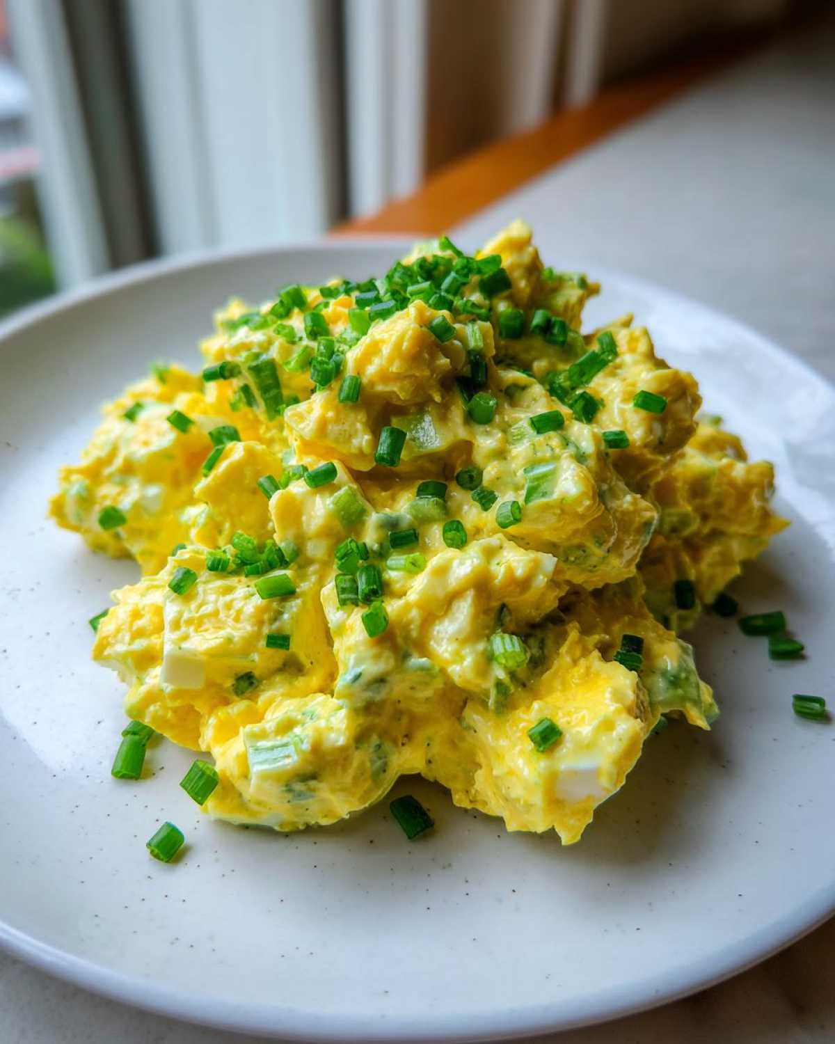 A mound of creamy, yellow egg salad recipe garnished generously with freshly chopped green chives on a white plate.
