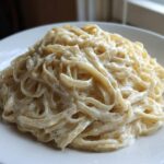 A close-up of fettuccine pasta generously coated in rich, creamy homemade alfredo sauce, served on a white plate.