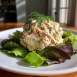 A beautiful scoop of creamy salmon salad mixed with celery, served on a bed of mixed greens and topped with fresh dill.
