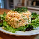 A perfectly scooped portion of creamy salmon salad served on a bed of fresh green and red lettuce, garnished with dill.