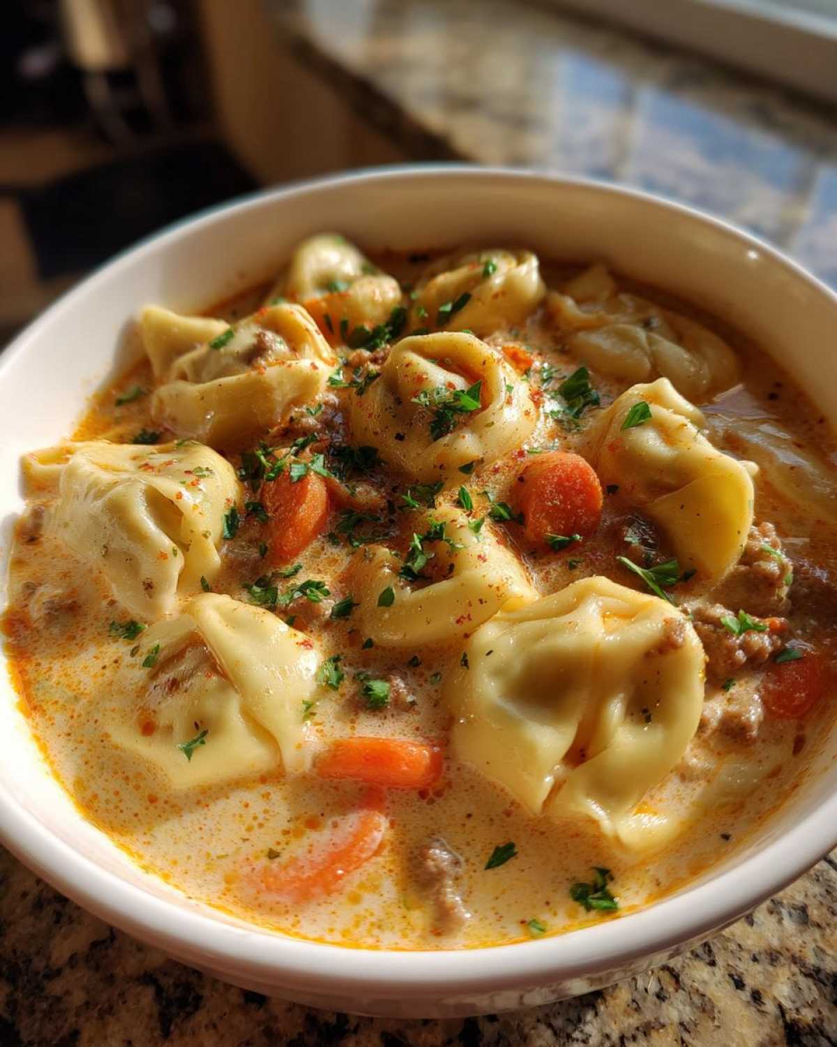 Close-up of a white bowl filled with creamy tortellini soup, featuring cheese tortellini, ground meat, sliced carrots, and parsley.