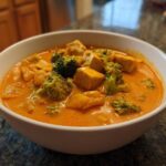A white bowl filled with vibrant orange creamy vegan pumpkin curry featuring chunks of tofu and bright green broccoli florets.