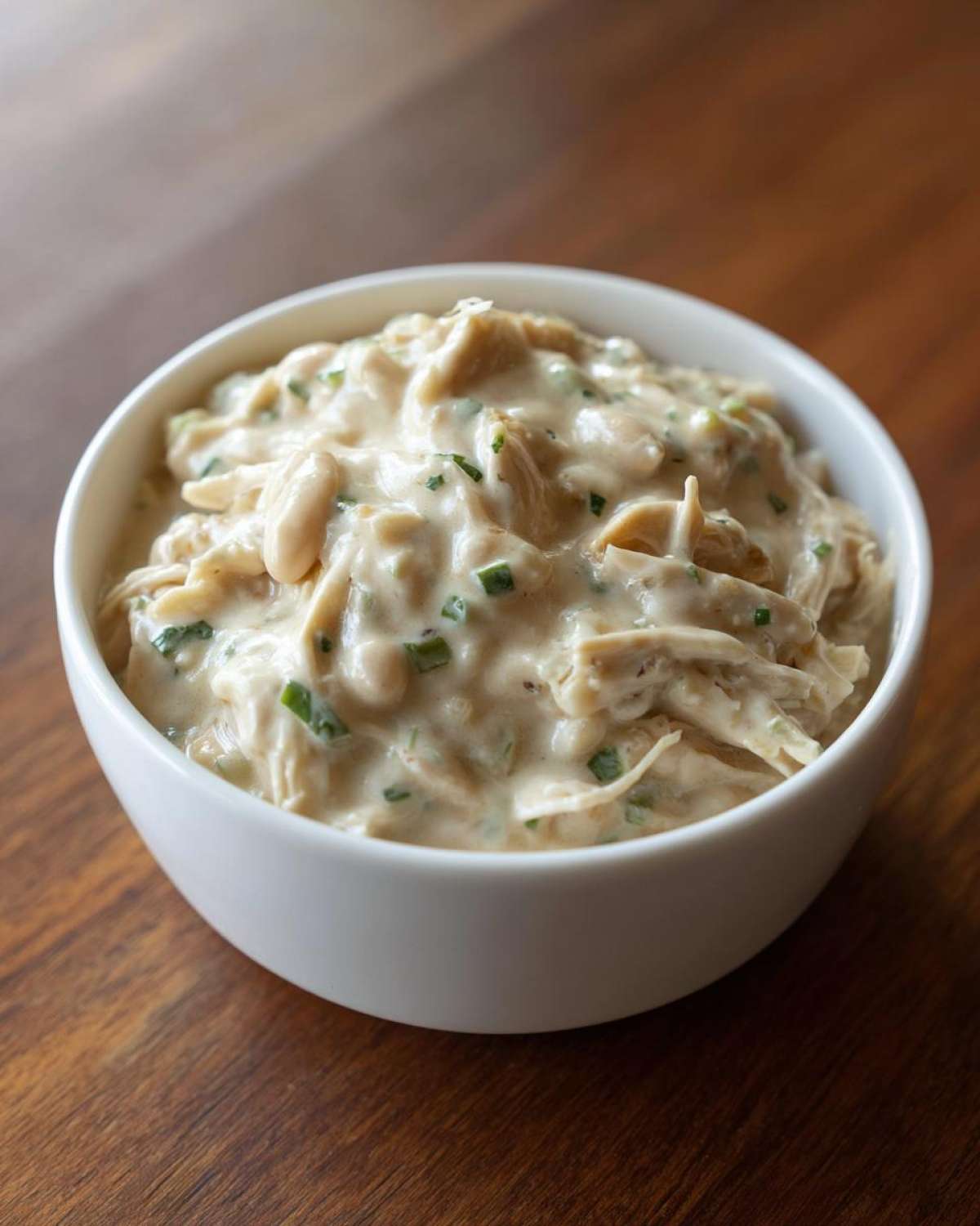 A white bowl filled with creamy white chicken chili recipe mixture, featuring shredded chicken and beans, topped with green herbs.