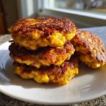 A stack of three golden brown, crispy corn fritters with one resting beside them on a white plate.