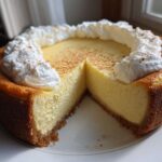 A whole eggnog cheesecake recipe with a slice removed, topped with whipped cream swirls and cinnamon.