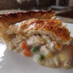 Close-up of a slice of turkey pot pie showing flaky golden crust and creamy filling with turkey chunks, peas, and carrots.