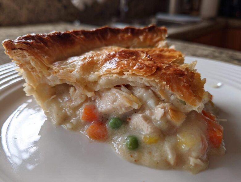 Close-up of a slice of turkey pot pie showing flaky golden crust and creamy filling with turkey chunks, peas, and carrots.