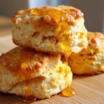 Three fluffy sweet potato biscuits stacked and drizzled generously with golden honey on a wooden surface.